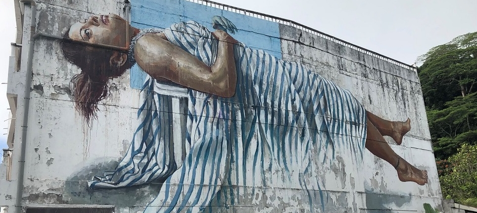 Painting on side of building on Raiatea. It's a woman laying on her back who appears to be floating, she's wearing a blue & shite thin striped dress.